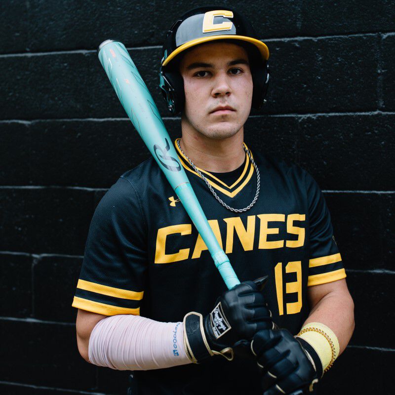 A baseball player in a black and yellow Canes uniform and cap, with headphones on and a bandaged left forearm, stands against a dark brick wall holding a 2024 Rawlings Mach AI (-3) BBCOR Baseball Bat: RBB4MC3 (USED).