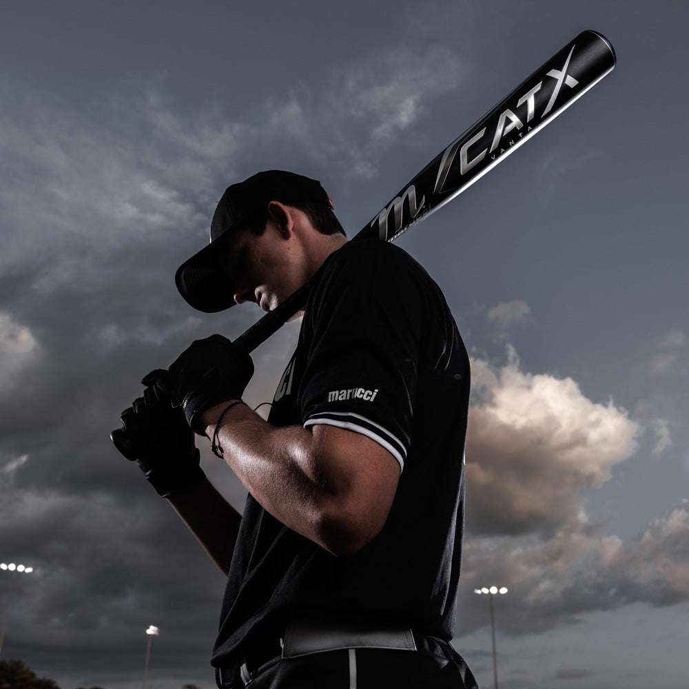 A baseball player in a black uniform and cap holds the Marucci 2024 CATX Vanta (-3) BBCOR Baseball Bat (MCBCXV) over his shoulder, standing outdoors under a cloudy evening sky with stadium lights behind him.