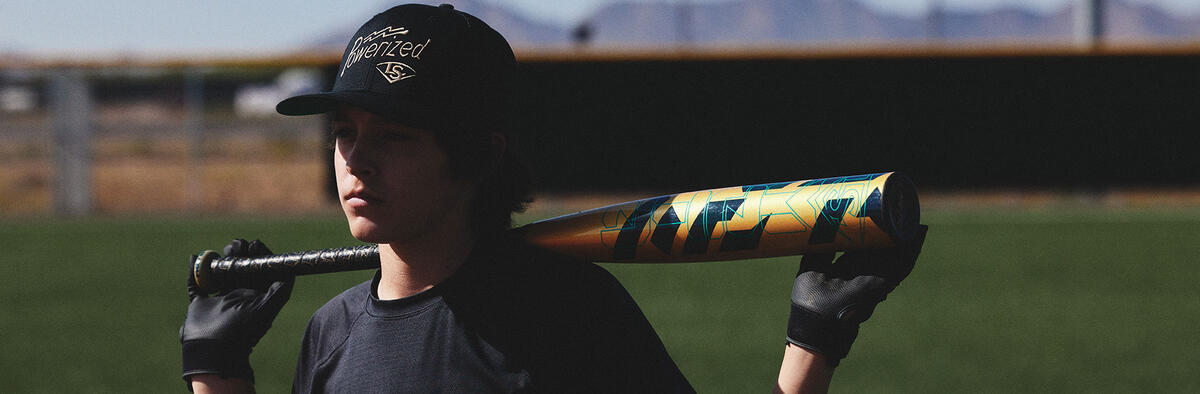 A young baseball player in a black shirt and cap stands on a field, holding a gold and blue bat across his shoulders. Mountains and a fence are visible in the blurred background.