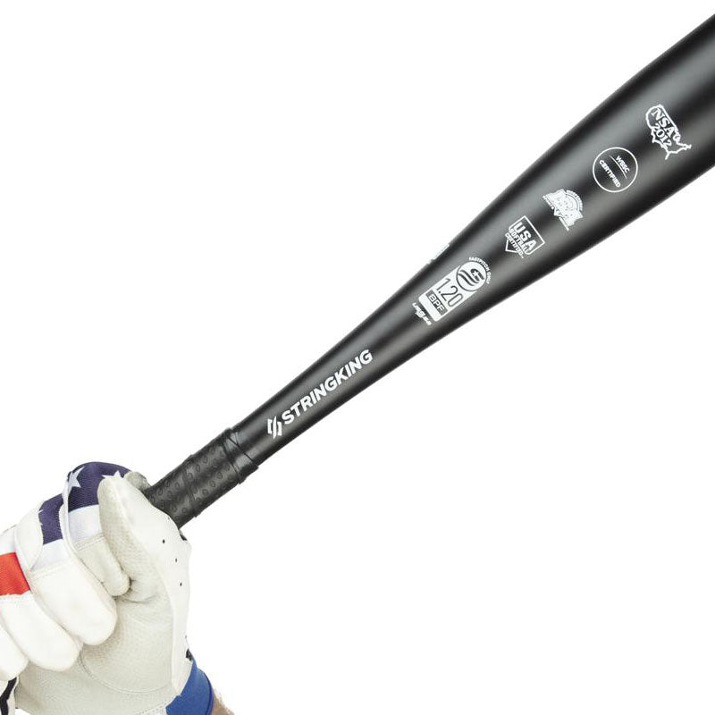 A gloved hand grips a black StringKing baseball bat, which features various certification logos printed in white near the handle on a white background.