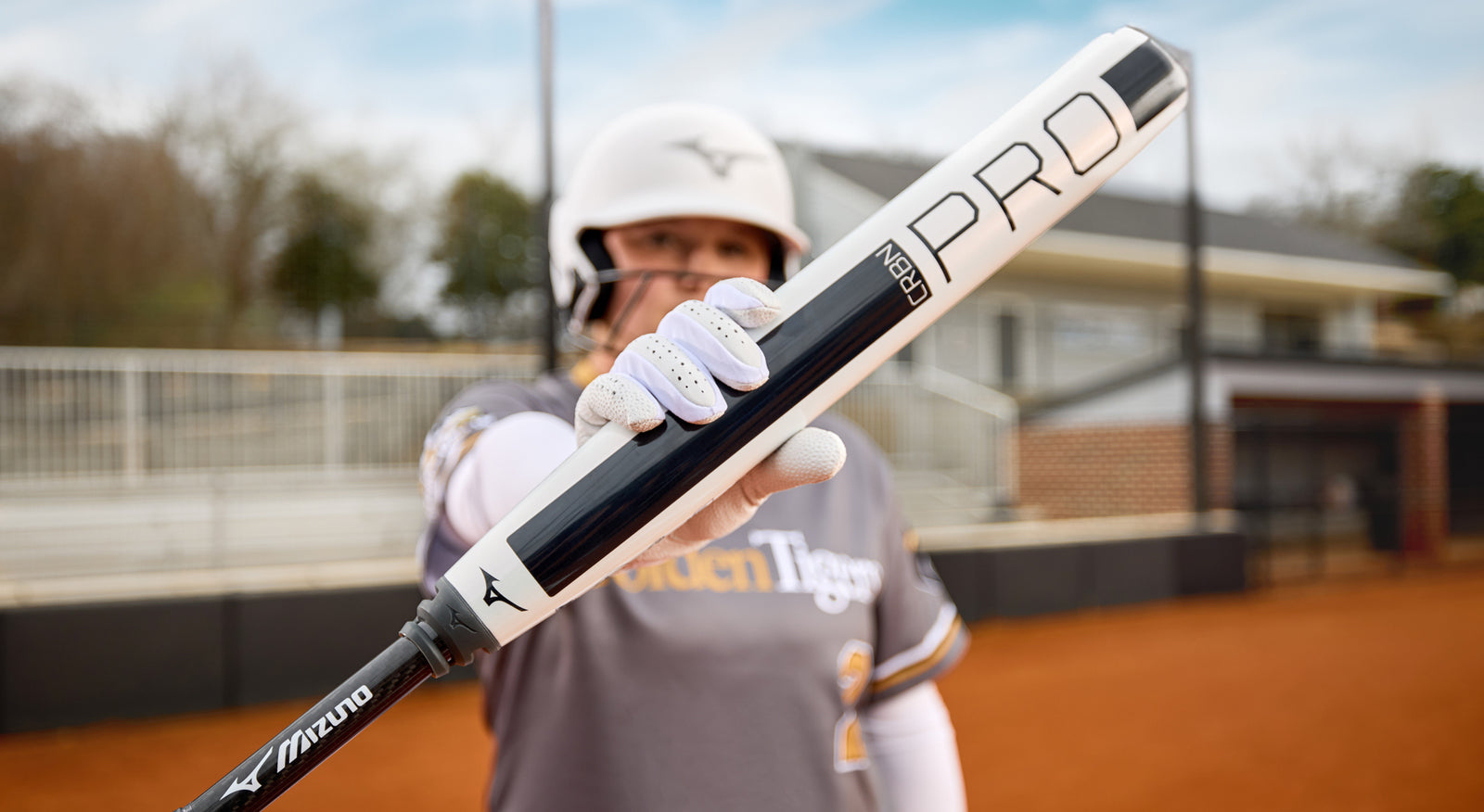 A softball player in a helmet and uniform stands on a field holding a Mizuno Pro bat toward the camera, with the bat as the main focus and the player blurred in the background.
