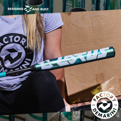A person in a gray DeMarini Factory tee holds a teal 2026 DeMarini Lotus (-11) Fastpitch Softball Bat (WBD2605010 DEMO) in front of a cardboard box. DeMarini logos appear on the shirt and in the corner of the image.