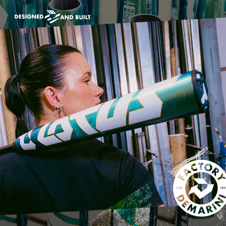 A woman with dark hair tied back holds a DeMarini 2026 Lotus (-11) Fastpitch Softball Bat (WBD2605010 DEMO) over her shoulder in front of metal pipes. DeMarini and Factory Designed and Built logos are overlaid on the image.