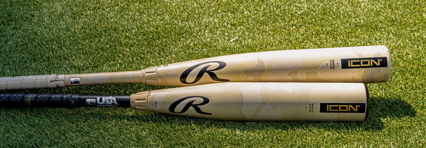 Two golden Rawlings ICON baseball bats with black handles and branding lie side by side on green grass in sunlight.