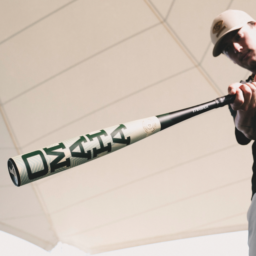 A baseball player in a cap showcases the Louisville Slugger Omaha (-3) BBCOR Baseball Bat (2026, WBL4117010), emphasizing its bold branding and one-piece alloy construction under a bright, white geometric canopy.
