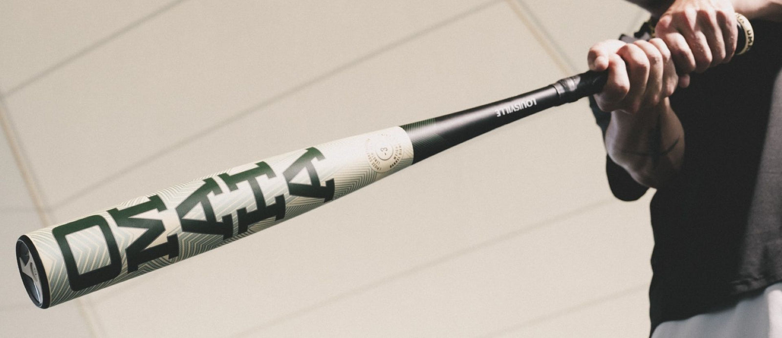A person holds a baseball bat with both hands, extending it outward. The bat has a bold, modern design with green and white patterns. The background is out of focus, highlighting the bat.