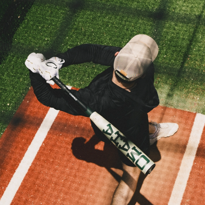 Overhead view of a person in a cap and gloves ready to swing the 2026 Louisville Slugger Omaha (-3) BBCOR Baseball Bat (WBL4117010) on a batting mat, surrounded by green artificial turf.