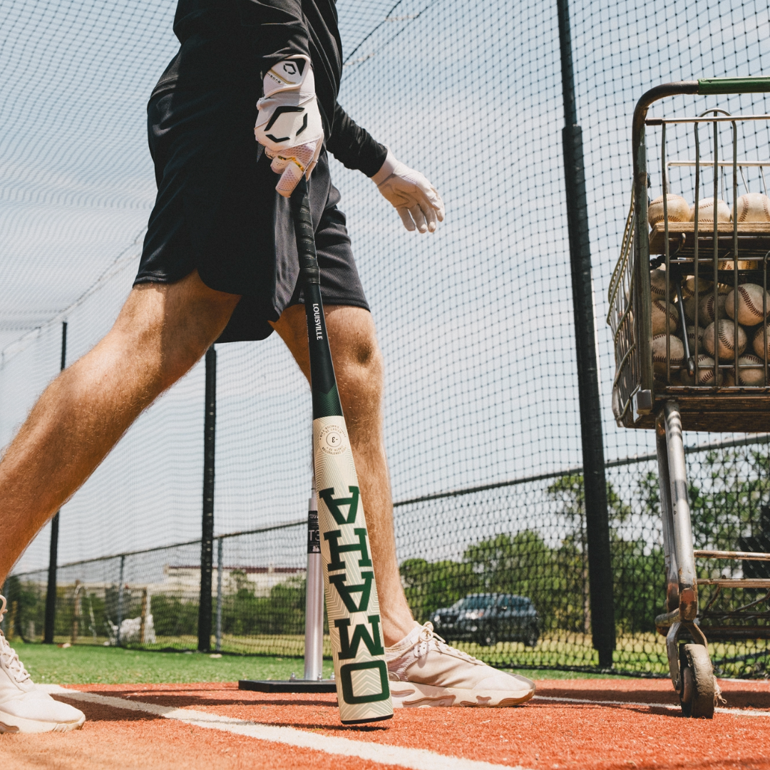 Wearing a glove, a person holds the 2026 Louisville Slugger Omaha (-3) BBCOR Baseball Bat (WBL4117010) in a batting cage, with a wheeled baseball basket nearby and red and white mats underfoot, surrounded by netted fencing.