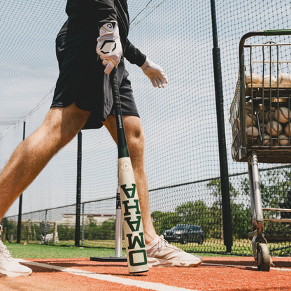 Wearing a glove, a person holds the 2026 Louisville Slugger Omaha (-3) BBCOR Baseball Bat (WBL4117010) in a batting cage, with a wheeled baseball basket nearby and red and white mats underfoot, surrounded by netted fencing.