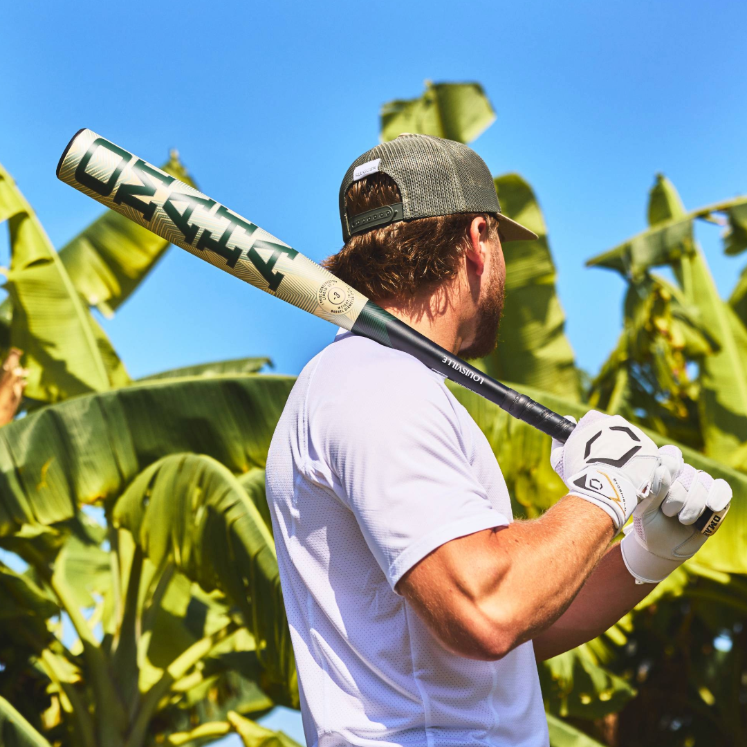 A man in a gray cap, white gloves, and a white shirt stands outdoors holding the 2026 Louisville Slugger Omaha (-3) BBCOR Baseball Bat (WBL4117010) over his shoulder with green banana leaves and blue sky behind him.