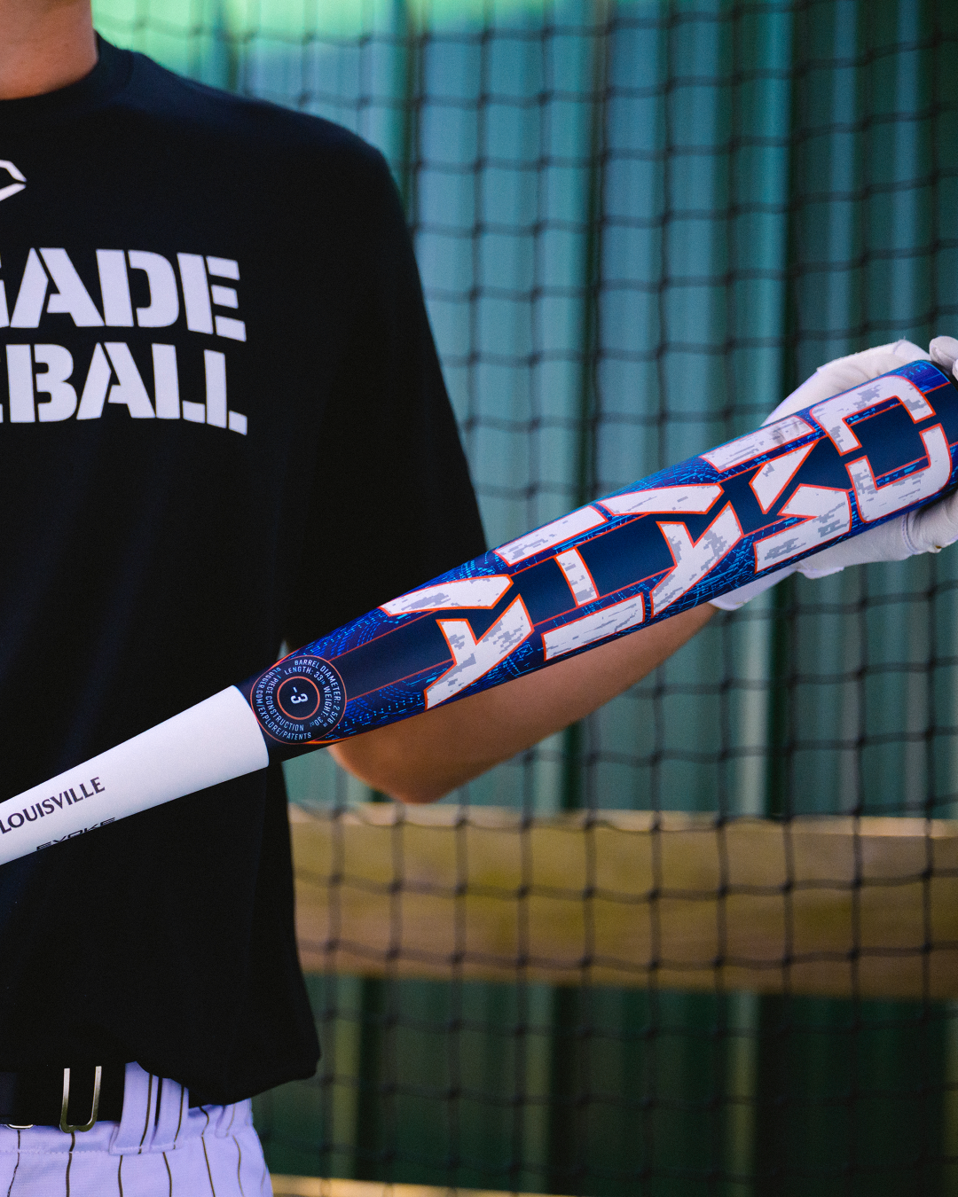 Wearing a MADE BASEBALL shirt, a person holds the 2027 Louisville Slugger Omaha (-3) BBCOR Baseball Bat (model WBL4249010), which is adorned with vibrant blue, red, and white geometric patterns, while standing in front of a netted background.