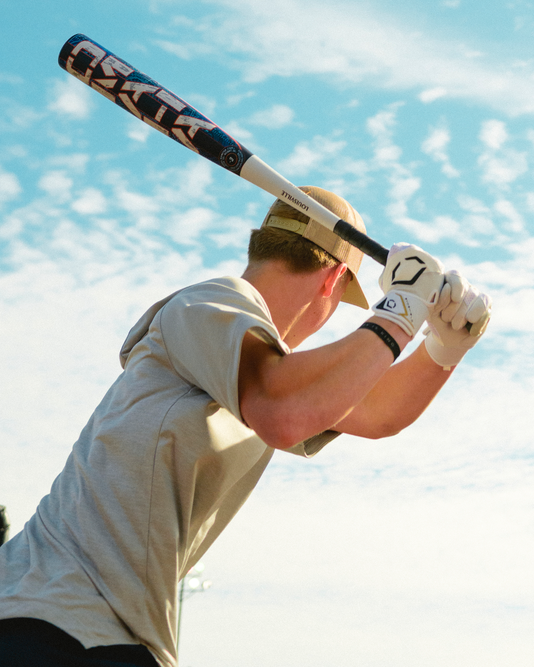A person wearing a hat and gloves prepares to swing the 2027 Louisville Slugger Omaha (-3) BBCOR Baseball Bat (WBL4249010) under a blue sky with scattered clouds.
