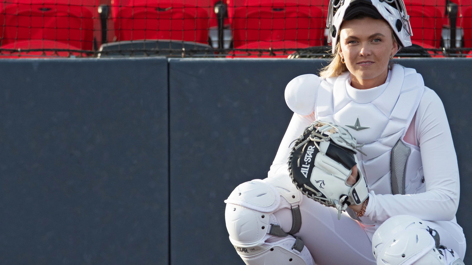 A softball catcher dressed in white protective gear crouches on the field, holding a glove, with empty red stadium seats in the background.