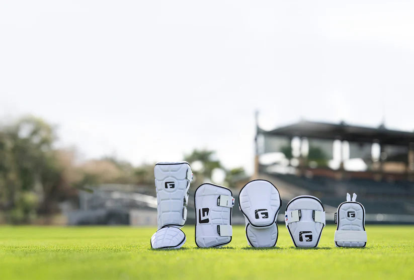 Five G-Form Alpha Batter's Elbow Guards (AEP132) by G-Form, designed for impact protection, are neatly arranged on green grass with a blurred stadium and trees in the background under a bright sky.