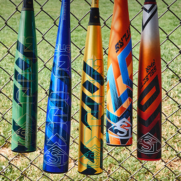 Five colorful baseball bats—green, blue, yellow, orange, and red—hang vertically on a chain-link fence with grass visible in the background.