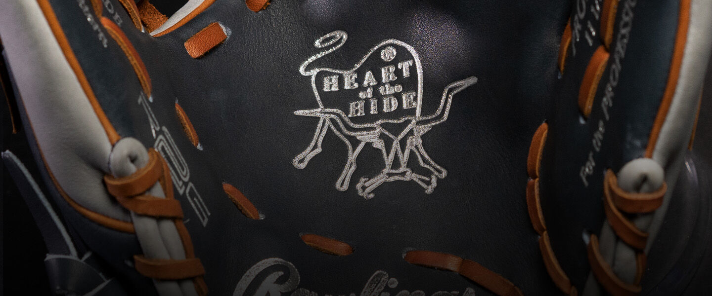 Close-up of a black baseball glove with tan laces. The inside features a stitched design of a cow and the words HEART of the HIDE in white thread. The Rawlings logo is partially visible at the bottom.