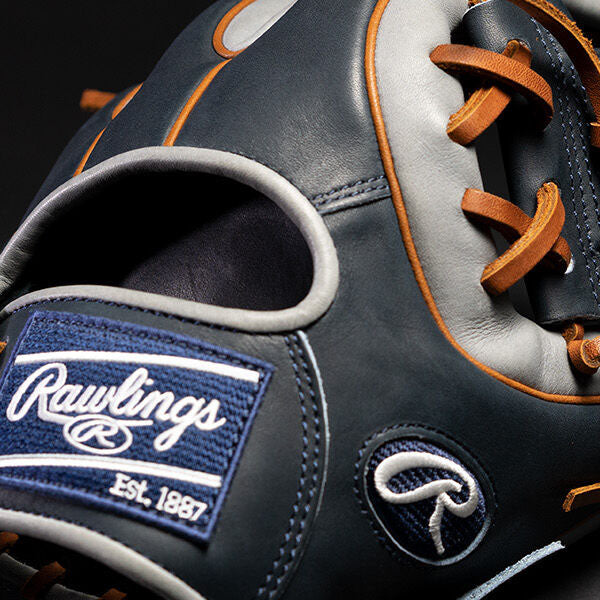 Close-up of a dark blue and gray Rawlings baseball glove with tan laces. The Rawlings logo patch and Est. 1887 are visible on the glove, which is set against a dark background.
