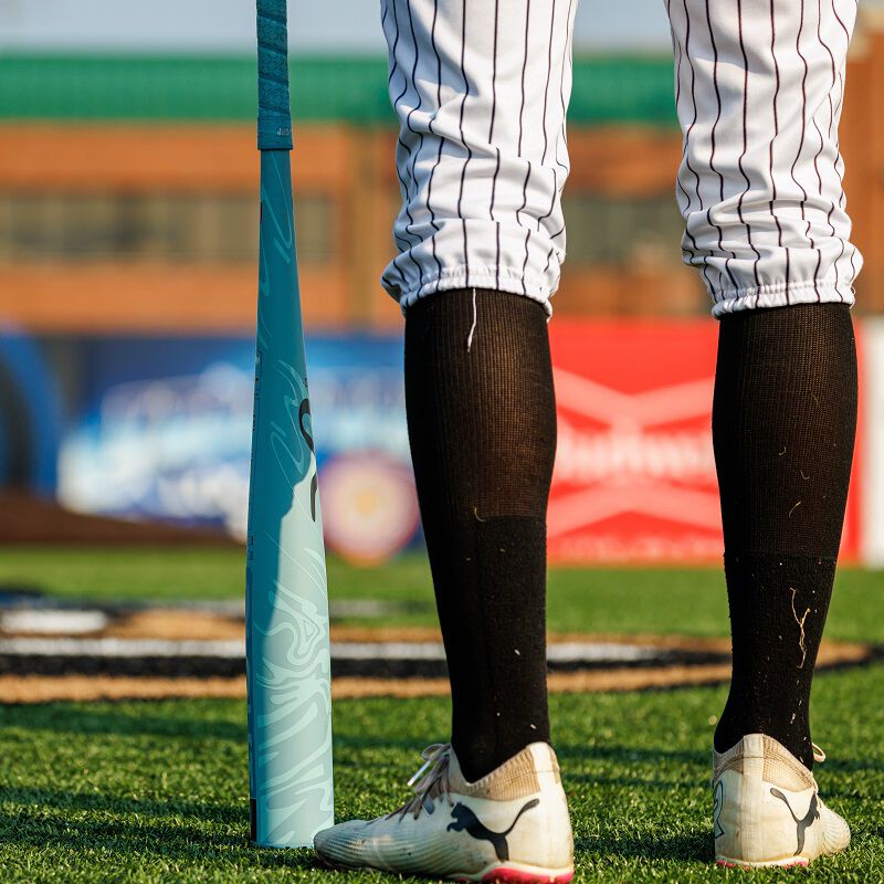 A baseball player in white cleats, black socks, and pinstriped pants stands on a field next to a 2025 Rawlings Clout AI (-3) BBCOR Baseball Bat: RBB5C3 (USED), with blurred stadium signage in the background.