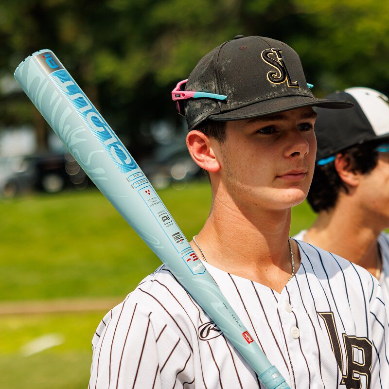 A young baseball player in a white pinstriped uniform and black cap holds a blue 2025 Rawlings Clout AI (-3) BBCOR Baseball Bat: RBB5C3 (USED) over his shoulder. Another player stands nearby, set against a green, blurry background.
