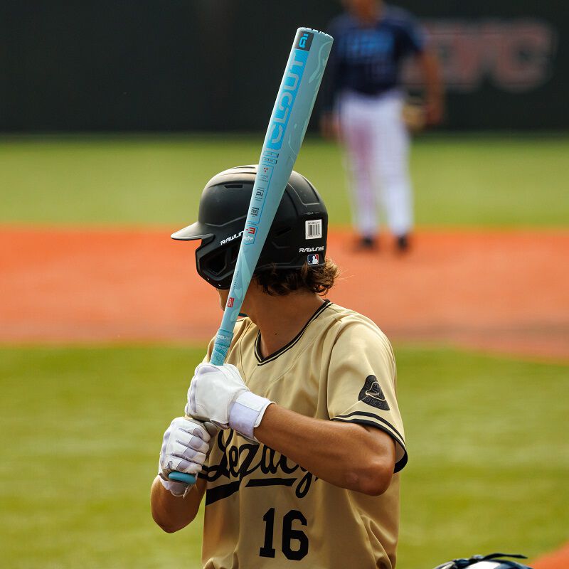 A baseball player in a gold jersey and black helmet grips a 2025 Rawlings Clout AI (-3) BBCOR Baseball Bat: RBB5C3 (USED), ready to hit, while another player in a blue uniform appears blurred in the background on the field.
