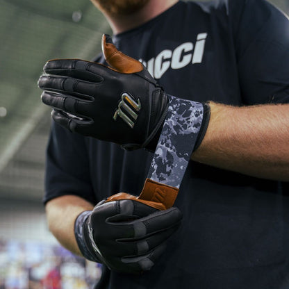 A person wearing black and brown Marucci REMX Youth Batting Gloves (MBRMXY) with the M logo adjusts the strap on their opposite wrist. The cabretta sheepskin glove is paired with a dark shirt featuring partially visible white lettering.