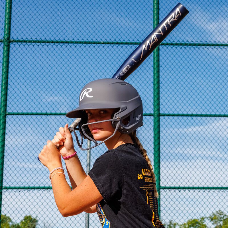 A young softball player, wearing a helmet, grips a 2025 Rawlings Mantra (-9) Fastpitch Softball Bat: RFP4M9 (DEMO) at the plate on a sunny day. A green chain-link fence and blue sky fill the background. Brand: Rawlings.
