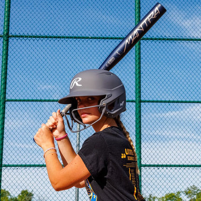 A young softball player, wearing a helmet, grips a 2025 Rawlings Mantra (-9) Fastpitch Softball Bat: RFP4M9 (DEMO) at the plate on a sunny day. A green chain-link fence and blue sky fill the background. Brand: Rawlings.