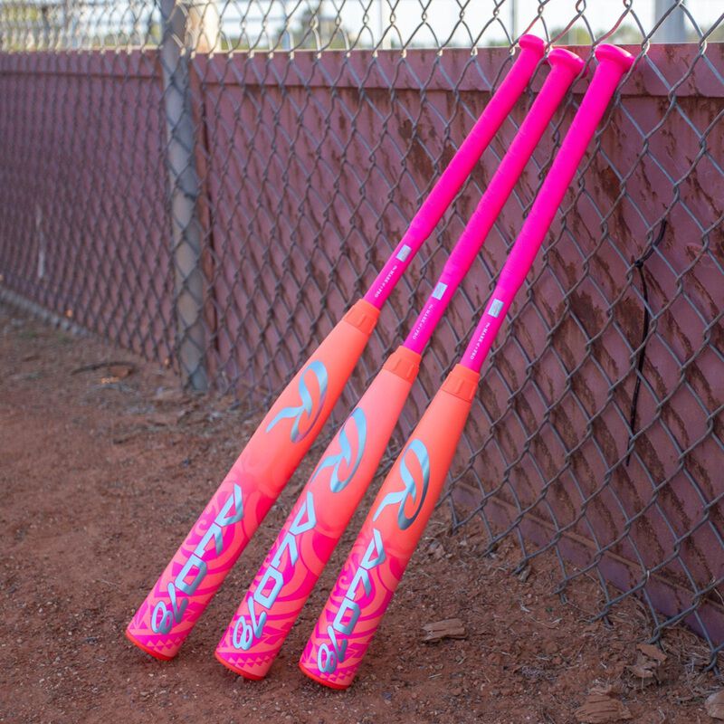 Three vibrant pink and orange Rawlings 2026 ALO78 (-12) Fastpitch Softball Bats with bold white logos rest against a chain-link fence on a dirt field.