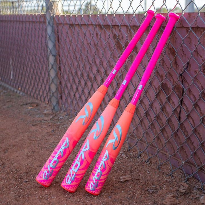 Three vibrant pink and orange Rawlings 2026 ALO78 (-12) Fastpitch Softball Bats with bold white logos rest against a chain-link fence on a dirt field.