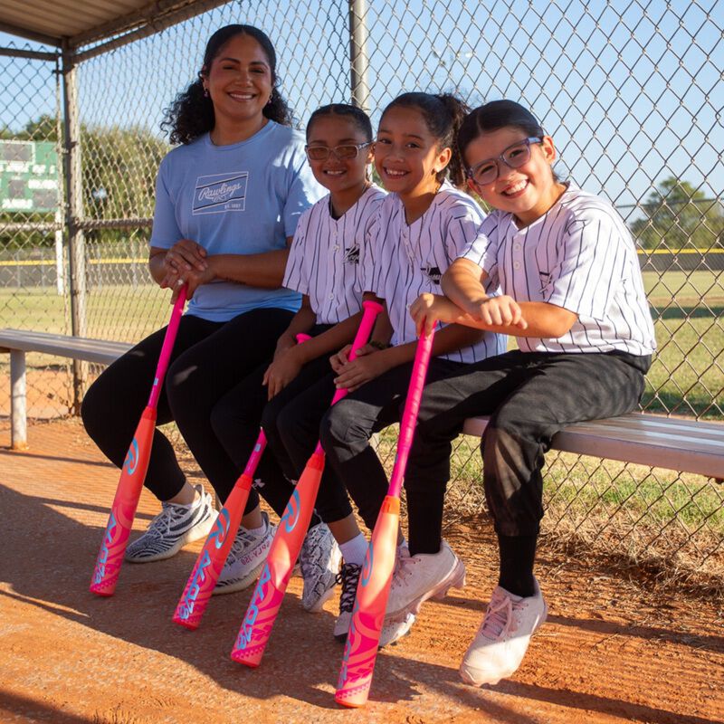 A woman and three smiling girls in baseball uniforms sit on a bench, each holding a 2026 Rawlings ALO78 (-12) Fastpitch Softball Bat: RFP6ALO12, with the sunny softball field in the background.