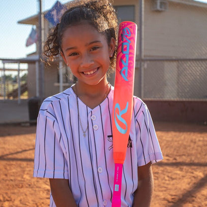 A smiling girl in a striped baseball jersey stands on a ball field, holding a vibrant Rawlings 2026 ALO78 (-12) Fastpitch Softball Bat (RFP6ALO12), with a chain-link fence and building visible behind her.