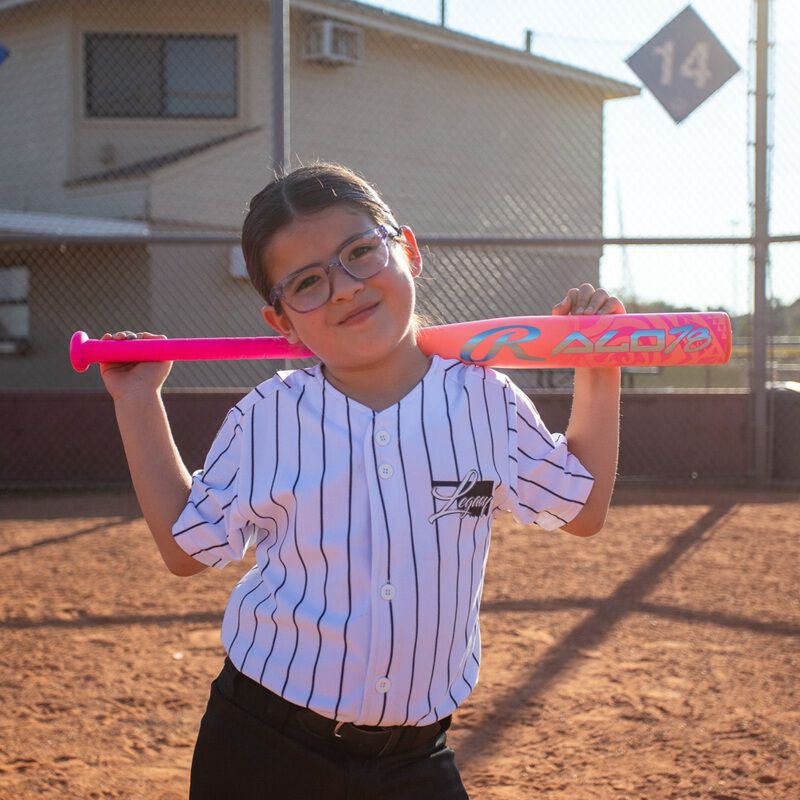 A young girl in glasses and a striped baseball jersey stands on a baseball field, smiling at the camera as she holds a bright pink Rawlings 2026 ALO78 (-12) Fastpitch Softball Bat (RFP6ALO12) with intense carbon construction across her shoulders.