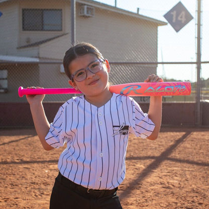 A young girl in glasses and a striped baseball jersey stands on a baseball field, smiling at the camera as she holds a bright pink Rawlings 2026 ALO78 (-12) Fastpitch Softball Bat (RFP6ALO12) with intense carbon construction across her shoulders.