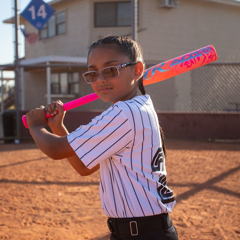 A young girl in a striped uniform and glasses stands ready to swing on a field, holding the Rawlings 2026 ALO78 (-12) Fastpitch Softball Bat (RFP6ALO12), with a building and a blue "14" sign visible in the background.