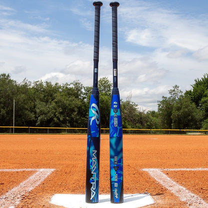 Two blue and black fastpitch bats, including the 2026 Rawlings Mantra (-11) Fastpitch Softball Bat: RFP6M11 (DEMO), stand upright on home plate of an orange dirt softball field, with trees and a cloudy sky in the background.