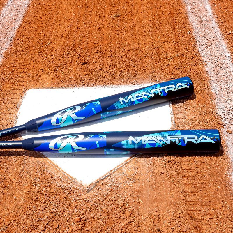 Two blue and black 2026 Rawlings Mantra (-11) Fastpitch Softball Bats (RFP6M11), made with In/Tense Carbon Composite, lie on home plate of a dirt softball field, with white chalk baselines in view.