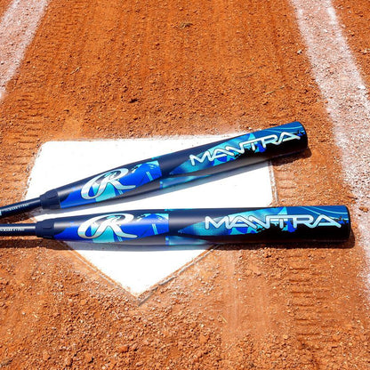 Two blue and black 2026 Rawlings Mantra (-11) Fastpitch Softball Bats (RFP6M11), made with In/Tense Carbon Composite, lie on home plate of a dirt softball field, with white chalk baselines in view.