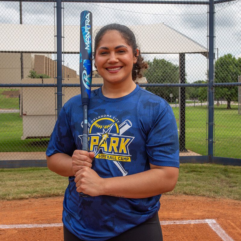 A smiling woman in a blue Park Softball Camp shirt holds the 2026 Rawlings Mantra (-11) Fastpitch Softball Bat (DEMO) on a softball field, with a fence and green grass in the background.
