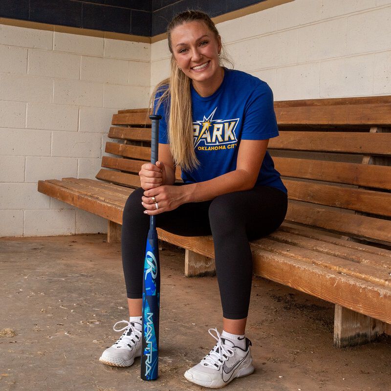 A smiling woman in athletic wear sits on a wooden bench in a dugout, holding the 2026 Rawlings Mantra (-11) Fastpitch Softball Bat (DEMO) vertically. She wears a blue Spark Oklahoma City shirt, black leggings, and white sneakers.