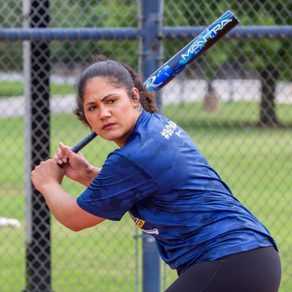 A woman in a blue sports shirt grips her Rawlings 2026 Mantra (-11) Fastpitch Softball Bat (RFP6M11 DEMO), poised to bat during a softball game, with a chain-link fence and trees visible in the background.