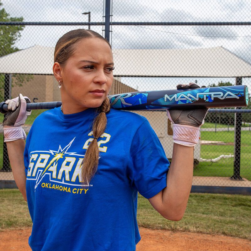 A softball player in a blue Spark Oklahoma City shirt stands on a field with a fence and dugout behind her, holding a 2026 Rawlings Mantra (-11) Fastpitch Softball Bat: RFP6M11 (DEMO) on her shoulder and wearing batting gloves.