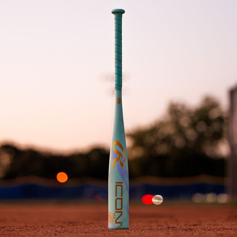 The 2026 Rawlings ICON CHOSEN ONE (-10) USA Baseball Bat (RUS6IONE10) stands upright on a dirt field at sunset, with blurred trees and lights in the background.