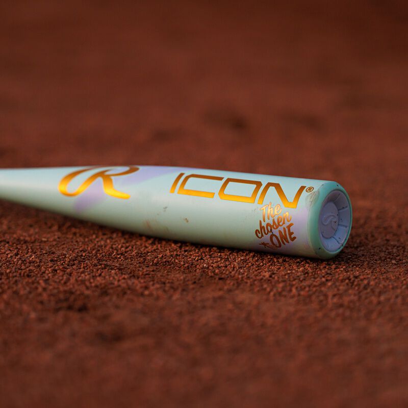 A close-up of the 2026 Rawlings ICON CHOSEN ONE (-10) 2 5/8" USA Baseball Bat shows its light green composite design with gold ICON and The Chosen One lettering, lying on reddish-brown dirt.