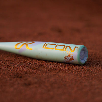 A close-up of the 2026 Rawlings ICON CHOSEN ONE (-10) 2 5/8" USA Baseball Bat shows its light green composite design with gold ICON and The Chosen One lettering, lying on reddish-brown dirt.