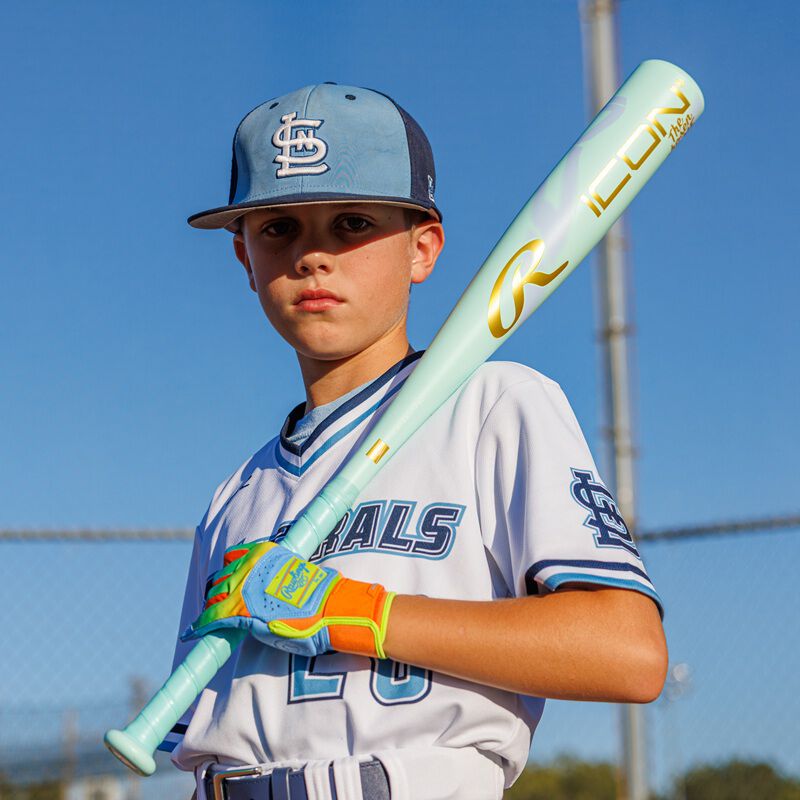 A young baseball player in a blue and white uniform holds the 2026 Rawlings ICON CHOSEN ONE (-10) USA Baseball Bat (RUS6IONE10) over his shoulder, sporting colorful gloves, with a chain-link fence and clear sky behind him.