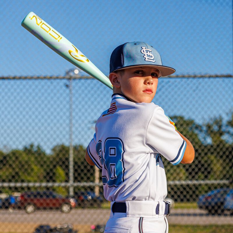 A young boy in a white baseball uniform and cap stands ready to bat on a sunny day, gripping his Rawlings 2026 ICON CHOSEN ONE (-10) USA Baseball Bat (RUS6IONE10). A chain-link fence and trees appear in the background.