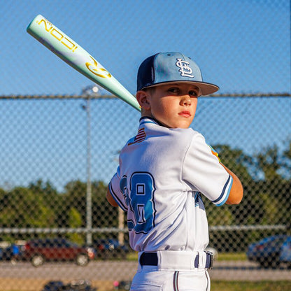 A young boy in a white baseball uniform and cap stands ready to bat on a sunny day, gripping his Rawlings 2026 ICON CHOSEN ONE (-10) USA Baseball Bat (RUS6IONE10). A chain-link fence and trees appear in the background.