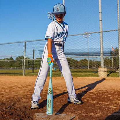 A young baseball player in a white and blue uniform stands in the batter’s box, holding the 2026 Rawlings ICON CHOSEN ONE (-10) USA Baseball Bat (RUS6IONE10), ready to hit, with a chain-link fence and grassy field behind on a sunny day.