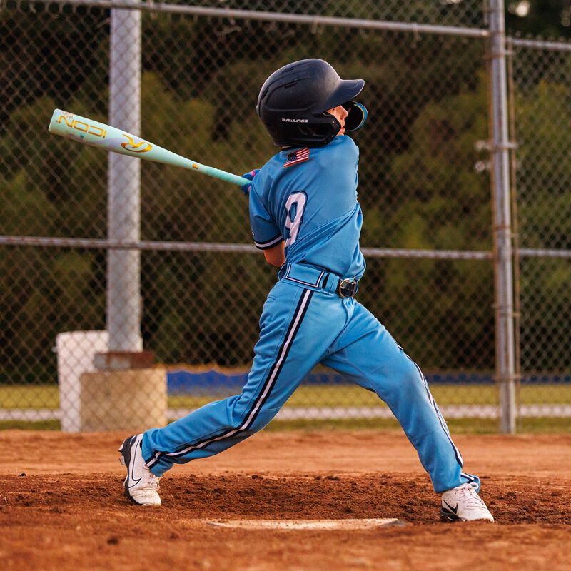 A young baseball player in a blue uniform swings a Rawlings 2026 ICON CHOSEN ONE (-10) 2 5/8" USA Baseball Bat (RUS6IONE10) on a dirt field, with a chain-link fence and green trees in the background.