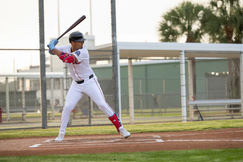 A baseball player in a white uniform with red accents stands ready to swing at home plate, wearing the G-Form Alpha Batter's Elbow Guard: AEP132 for superior impact protection on a fenced field with palm trees in the background.
